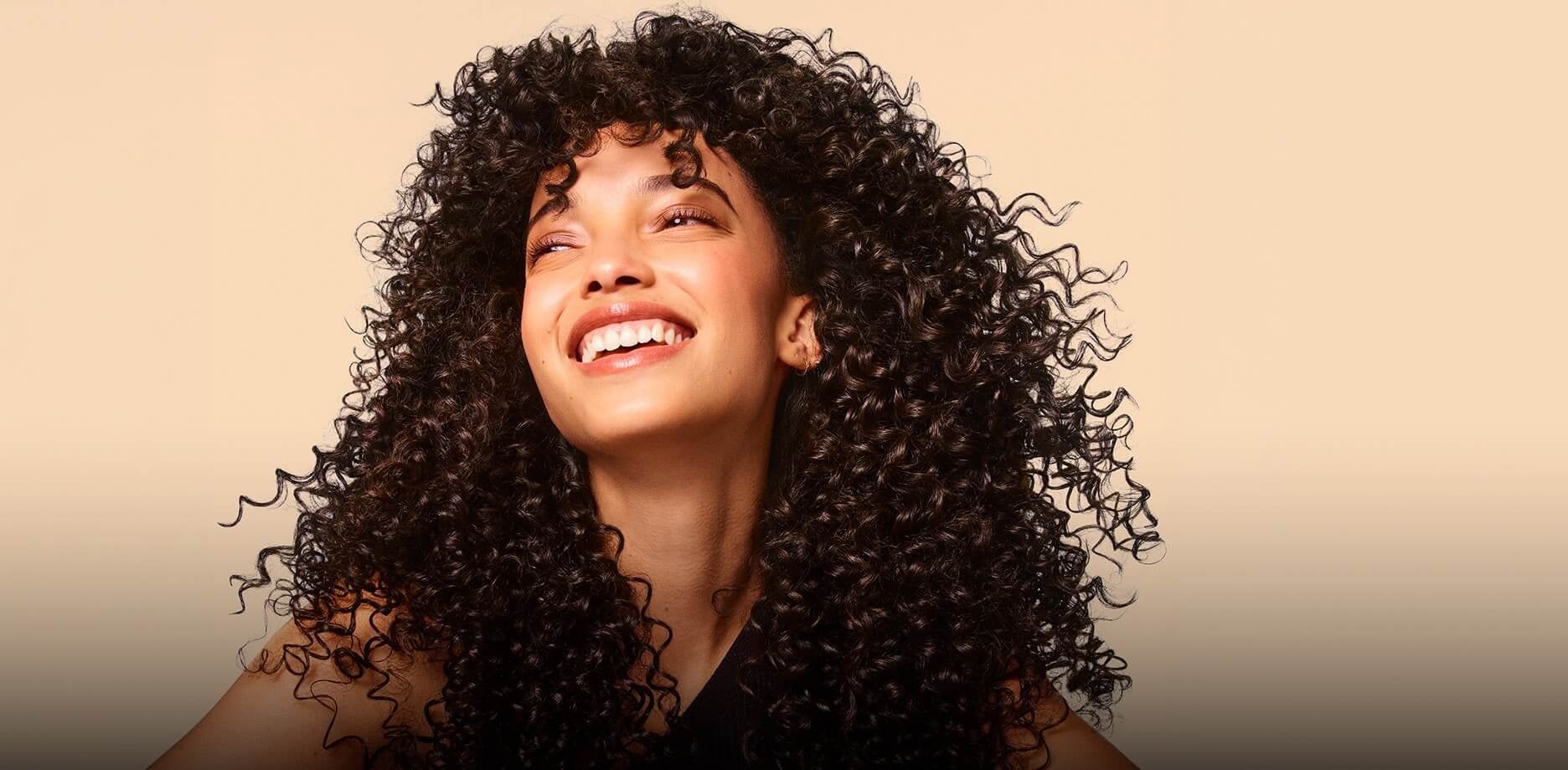 Woman with voluminous, tightly coiled curly dark hair smiling and looking upward against a beige background.