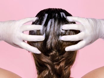 Gloved hands applying hair dye to the back of a brunette’s head.