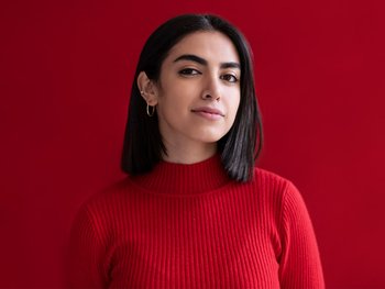 A woman with light skin and jet black hair poses against a red background.