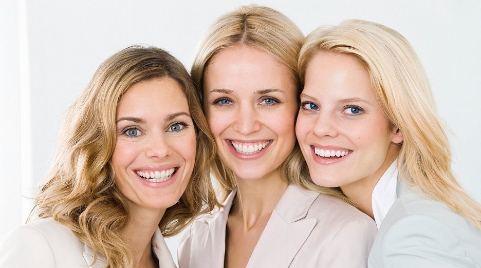 Three women with different shades of blonde hair smile at the camera, showcasing the variety of blonde hair colors.