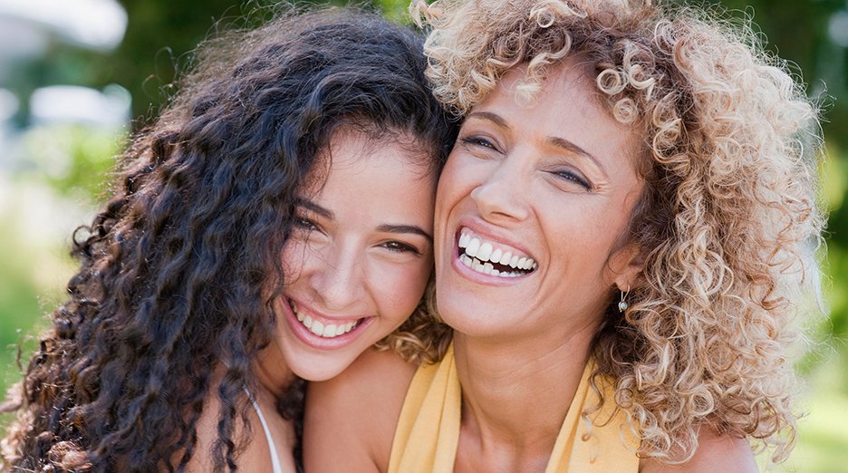 Close up of two female best friends laughing and embracing each other. They each have beautiful curly hair, one has long brunette 2C curls, while the other fashions shoulder length, blonde, 3B curls.