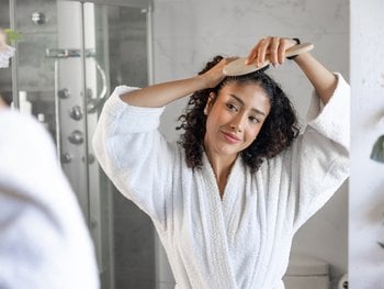 Young woman brushing curly hair in bathroom mirror.