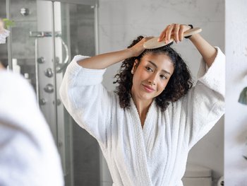 Young woman brushing curly hair in bathroom mirror.