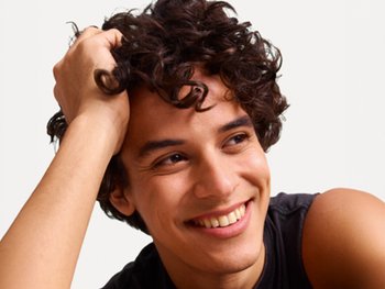 A young man with dark brown, tousled curly hair styled in defined ringlets smiles broadly while lifting a section of his hair with one hand, wearing a dark sleeveless top against a plain off-white background.