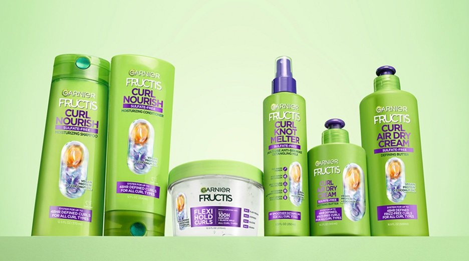 Garnier Fructis Curl haircare lineup displayed on a light green shelf against a matching green backdrop, featuring six green bottles and jars with purple accents and caps, including "Curl Nourish Shampoo," "Curl Nourish Conditioner," "Flexi Hold Curls" gel in a clear tub, "Curl Knot Melter" spray bottle, and "Curl Air Dry Cream”. Collection is made for all curl types and sulfate Free; a small caption at the bottom reads "*Packaging may vary."