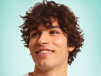Smiling young man with medium-length, loosely tousled dark curly hair against a mint green background.