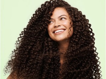 Close-up of a smiling woman with voluminous, tightly coiled dark curly hair framing her face against a beige background.