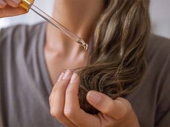 Close-up of a person applying hair oil or serum from a dropper onto the ends of their hair, demonstrating a nourishing hair treatment for dry or damaged strands.
