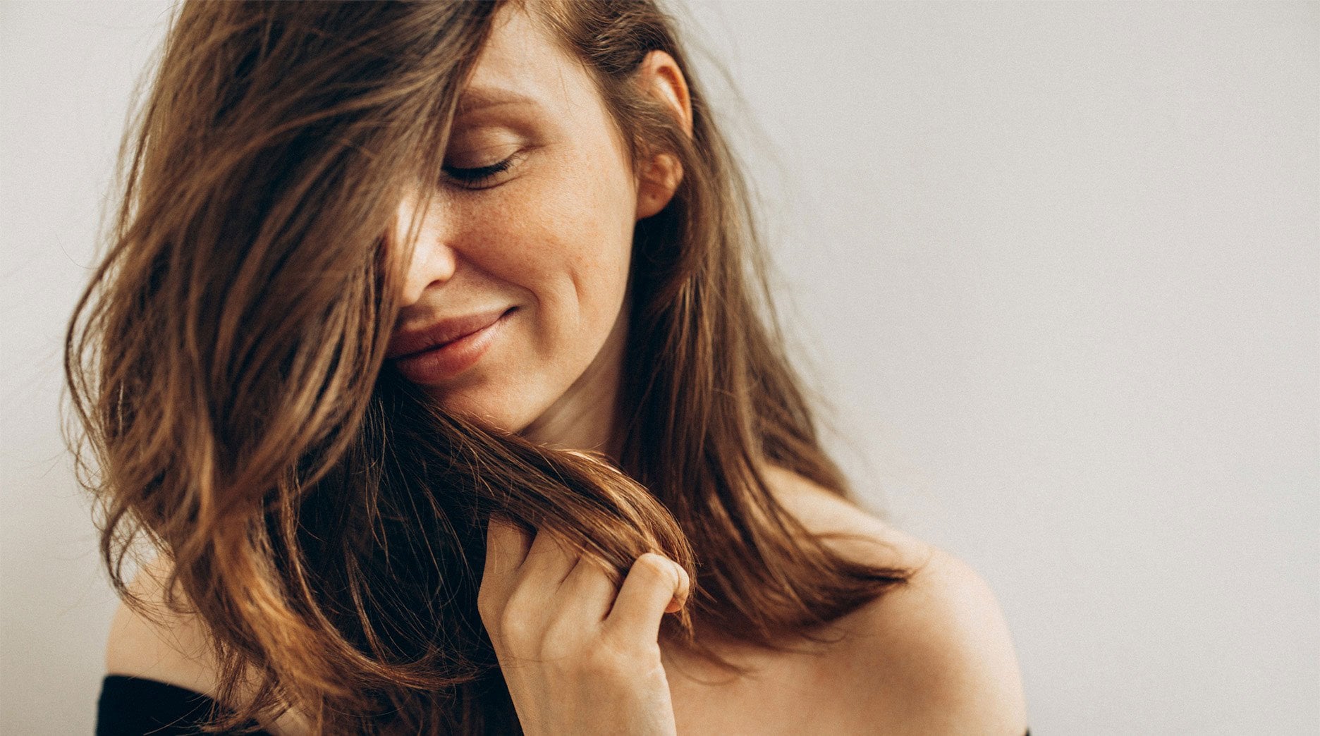 Beautiful woman with eyes closed as she hides half her face behind her smooth, wavy hair. She smiles embracing the softness.