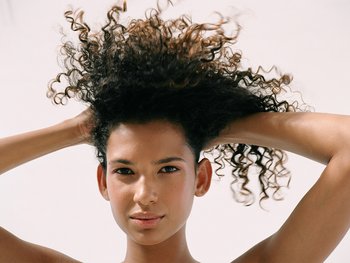 Woman with thick, curly hair, hands on her head.