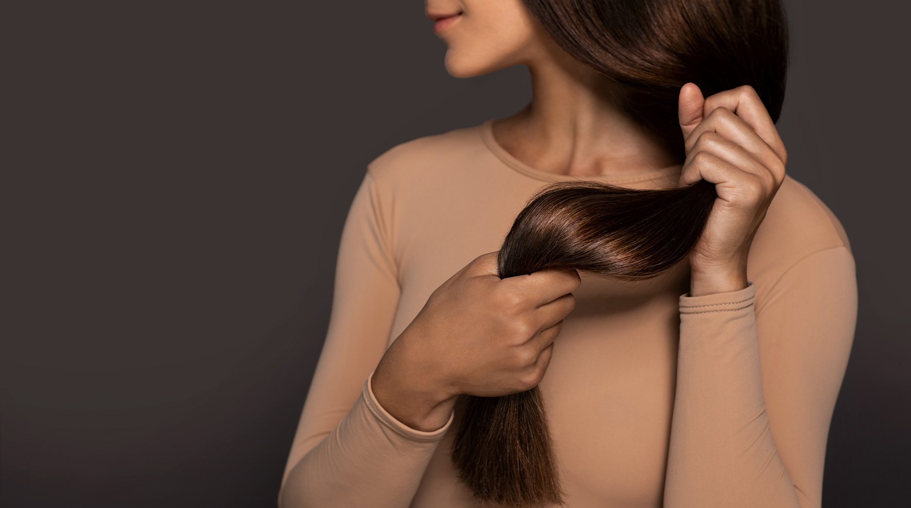 Brunette girl with long straight and shiny hair beauty skin woman holding her strong and healthy hair in her hands over grey background