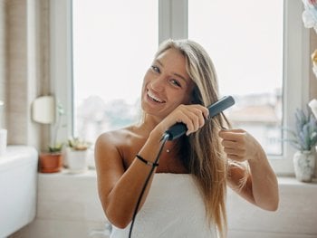 Blonde woman facing forward, smiling at camera as she straightens her long hair.