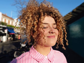 Young woman with 3B curls, curly hair styled in a half updo. She is smiling as she closes her eyes facing the sun.