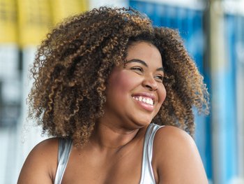 Young black woman with coily 4B curls smiling ash she looks to the side. Her hair is down, showcasing her beautiful defined curls.