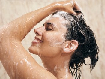 A woman with brown hair rinses her hair in the shower, clearly demonstrating the act of hair washing.