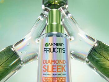 Garnier Fructis Diamond Sleek Shine-Coat Smoothing Spray bottle with a green pump cap is showcased amid glass-like leaf elements, promoting "100H Polished Shine, Frizz-Free Sleek."
