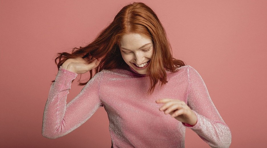 A woman with vibrant red hair smiles and runs her fingers through it, showcasing healthy and shiny hair.