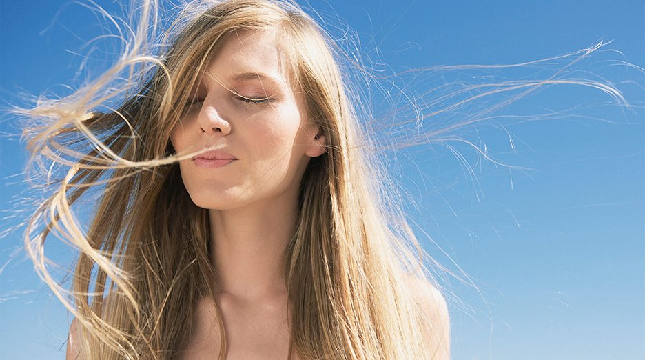 A person with long, light hair closes their eyes as their hair blows gently in the breeze.