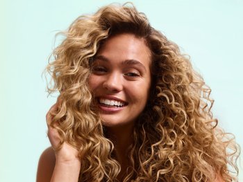 A smiling young woman with fair skin and voluminous, blonde-highlighted curly ringlets touches her defined curls with one hand against a mint-green background, showcasing Garnier Fructis curl care results.