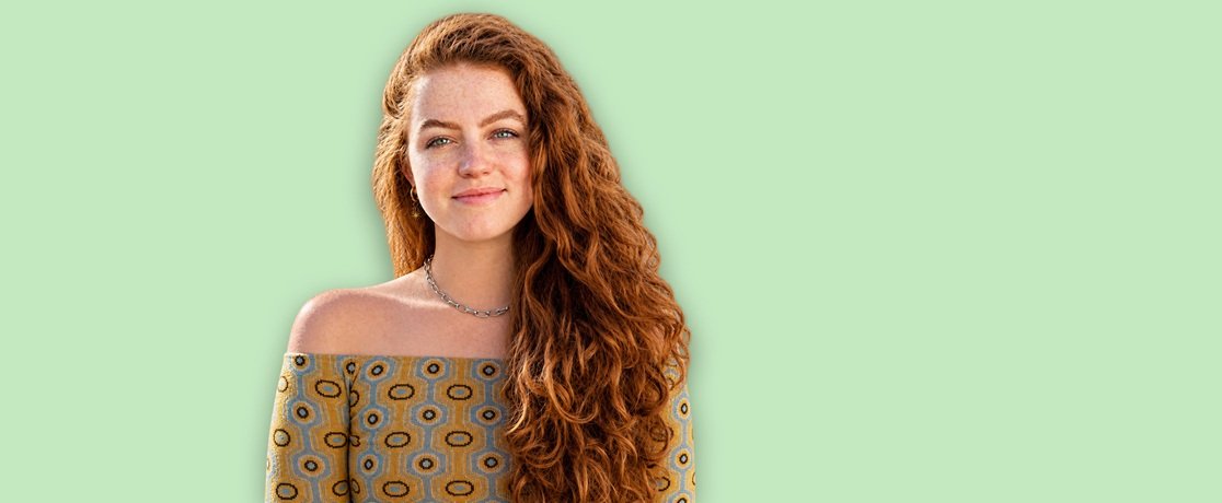 Young woman with fair, freckled skin and long, voluminous auburn-red curly hair with defined spiral curls falling over one shoulder, smiling softly in a silver chain necklace and an off-the-shoulder yellow, blue, and brown geometric-print top, showing curls styled with the Garnier Fructis Curl Nourish collection against a flat mint-green background. 