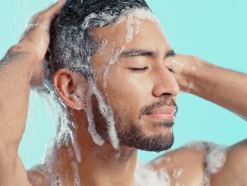 A person with short dark hair lathers their hair with shampoo.