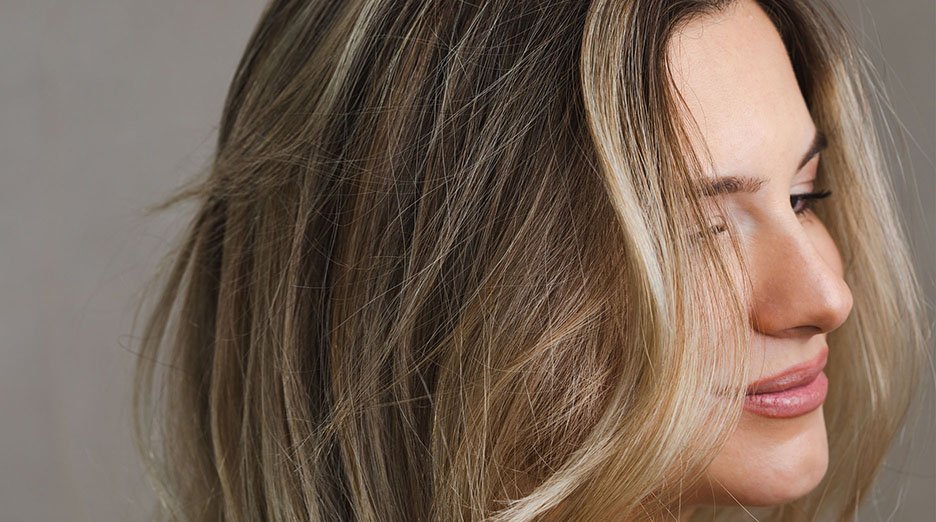 A close-up side profile of a woman with long, wavy blonde and brown highlighted hair, looking softly to the right, against a neutral gray background.