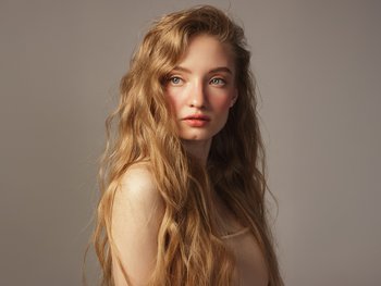 A young woman with long, wavy, golden blonde hair flowing over her shoulders, looking to the side with a soft expression against a grey background.