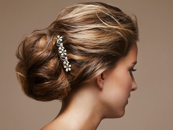 The profile of a woman with her blonde hair styled in an elegant updo, adorned with a decorative hair clip featuring light-colored stones, against a warm brown background.
