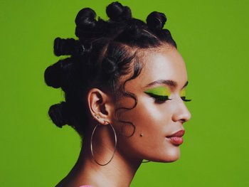 A woman with her hair styled in Bantu knots, wearing vibrant green eyeshadow and large hoop earrings, posing in profile against a bright green background.