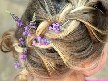 A close-up of a person's blonde hair styled in a decorative braid and bun, adorned with small purple flowers.