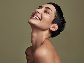 A person with short, dark hair and multiple piercings, laughing with their eyes closed and head tilted back, against a muted green background.