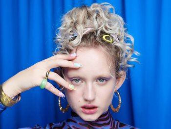 A woman with curly blonde hair styled with colorful hair clips, wearing bold makeup, chunky rings, and large hoop earrings, posing against a blue background.