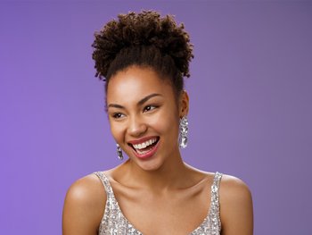 A smiling woman with dark, natural curly hair pulled up into a high puff, wearing a sequined top and large dangle earrings, looking joyfully to the side against a purple background.