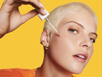 Model with platinum blonde pixie cut applying serum to face.