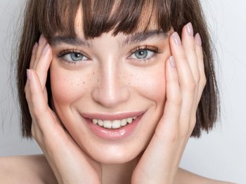Brunette model with blunt bob haircut holding her face while smiling at camera.