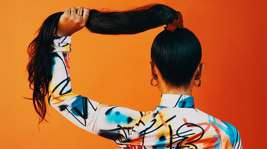 A person with long, dark hair holds their high ponytail against an orange background.