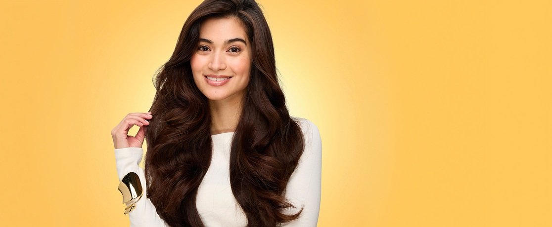 Smiling woman with voluminous, blown-out, glossy dark brown waves and caramel highlights after washing and drying with L'Oréal Honey Treasures shampoo, conditioner, and repairing serum, gently touching a silky strand of hair while wearing a cream top and gold cuff bracelet against a warm golden-yellow background.
