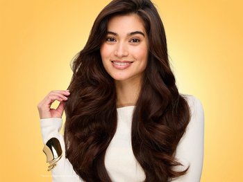 Smiling woman with voluminous, blown-out, glossy dark brown waves and caramel highlights after washing and drying with L'Oréal Honey Treasures shampoo, conditioner, and repairing serum, gently touching a silky strand of hair while wearing a cream top and gold cuff bracelet against a warm golden-yellow background.