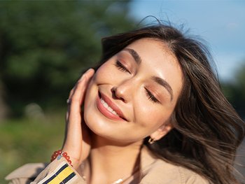 A smiling woman with closed eyes is enjoying the sunlight on her face outdoors, with natural light illuminating her features.