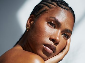 A close-up portrait of a person with intricate braided hair, looking intensely at the camera with their hand gently resting on their cheek, highlighting their radiant skin.