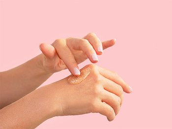 Two hands are shown, with one hand applying a light orange, granular scrub or product to the back of the other hand against a solid pink background.