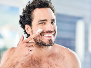 A smiling man applies moisturizer to his face, depicting the act of moisturizing.
