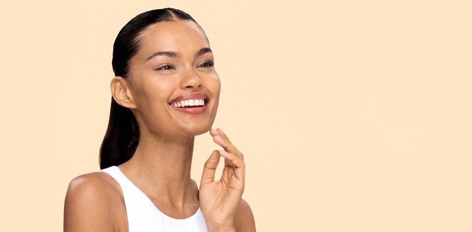A young woman with smooth, radiant tan skin and sleek dark hair pulled back smiles while touching her clear, hydrated chin, showcasing glowing skin achievable with micellar water cleansing, set against a soft peach background in a white tank top .