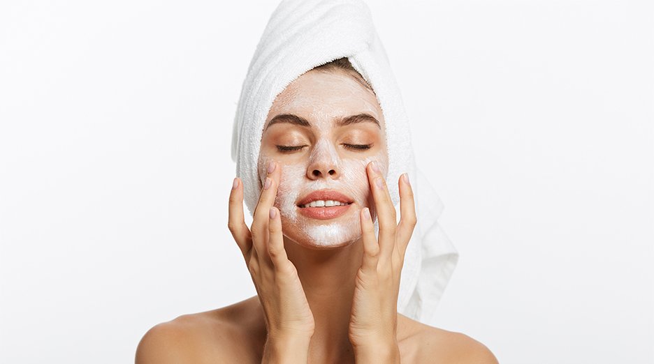 Close up of woman with hair wrapped in towel. Picture is from shoulders up, and she has her eyes closed as she uses her fingers to gently finish applying a cream mask to her recently cleansed face.