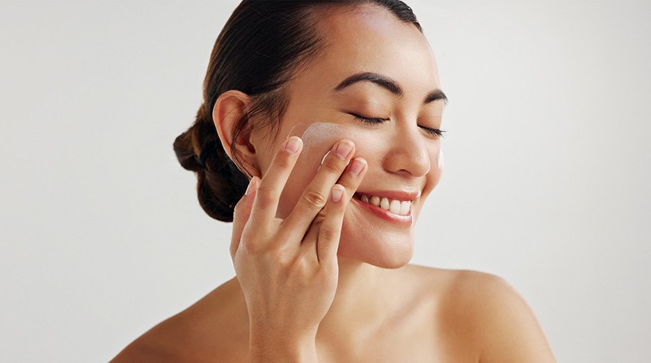 A smiling woman with her eyes closed, gently applying a white cream to her cheek with her fingers, against a light gray background.