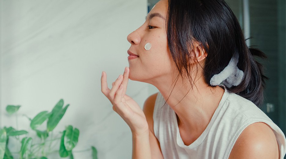 A close-up side profile of a person with dark hair, applying a skincare product to their face. A visible dollop of white cream is on their cheek, and their fingers are near their chin, ready to spread the product.