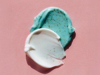 Two small mounds of scrub, one teal and one white, are shown on a pink background, representing exfoliating products.