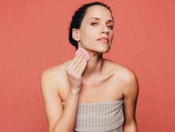 A woman applies cleanser to her neck with a cotton pad.