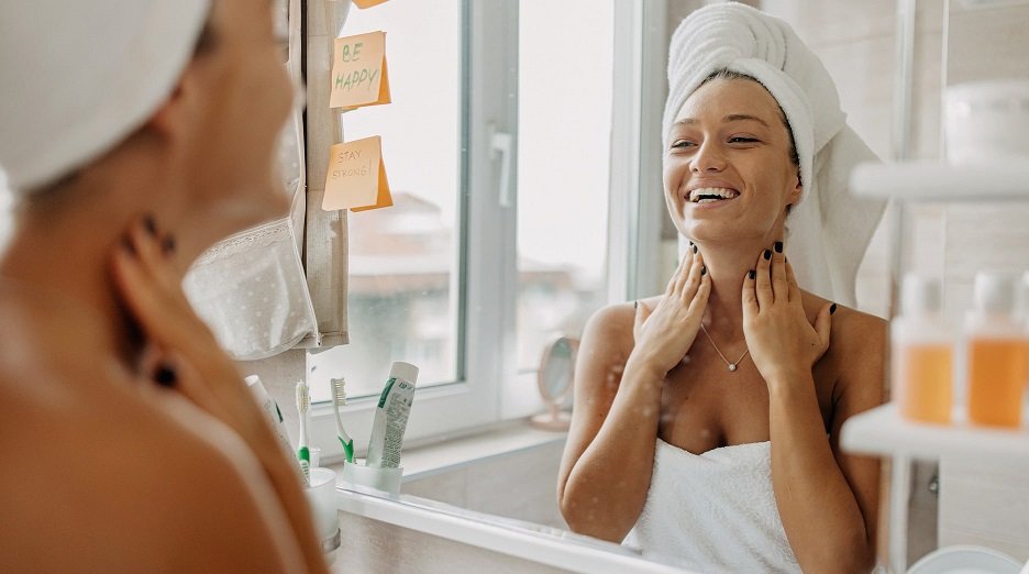 A smiling woman in a towel touches her neck while looking in the mirror, highlighting the importance of including the neck in skincare routines.
