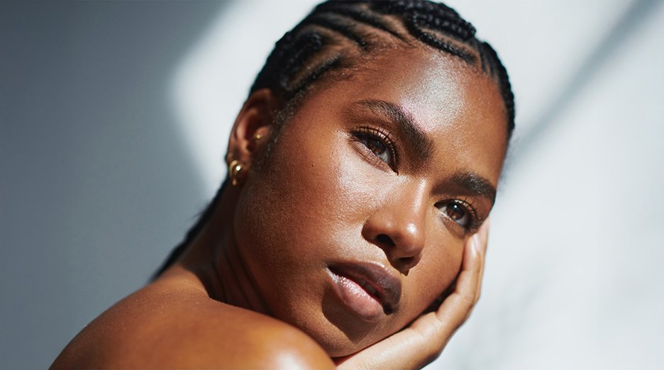 A person with dark skin, braided hair, and visibly oily skin leans their cheek on their hand.